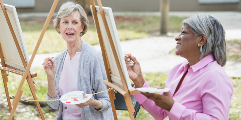 African american woman and caucasian woman painting outdoors at a life enrichment activity in a senior living community.