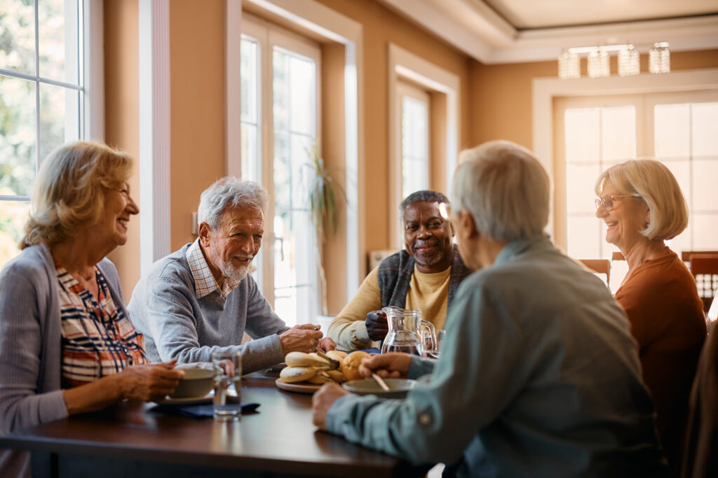 Residents smile at a dining table and enjoy bagels, fruit, and more for breakfast in a warm and inviting atmosphere.