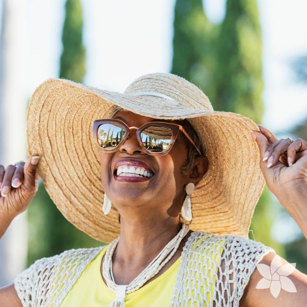 A woman outside, holding the rims of her floppy hat and smiling