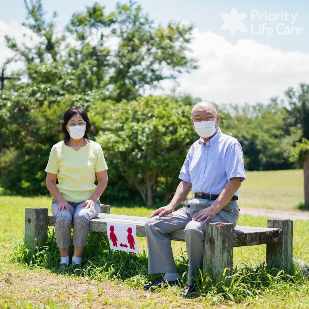 Senior couple outside wearing masks, sitting on a bench 5-feet apart.