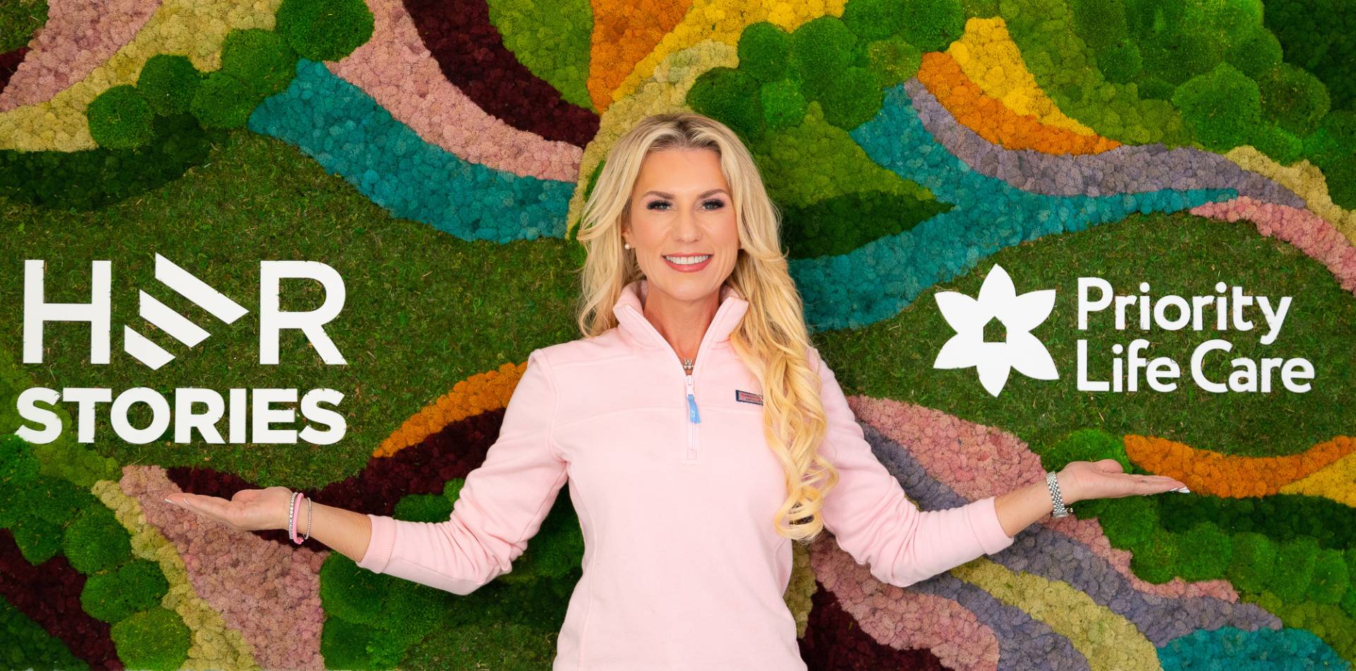 Sevy Petras standing in front of a wall that has the logos for both Her Stories and Priority Life Care